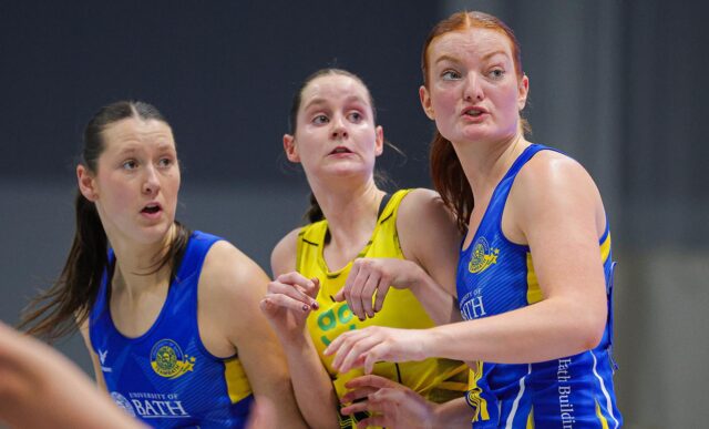 A photo of Team Bath Netball defenders Lottie Robinson and Scarlett Creasey closely marking a Manchester Thunder attacker during a 2025 NXT Gen League match in Bristol.
