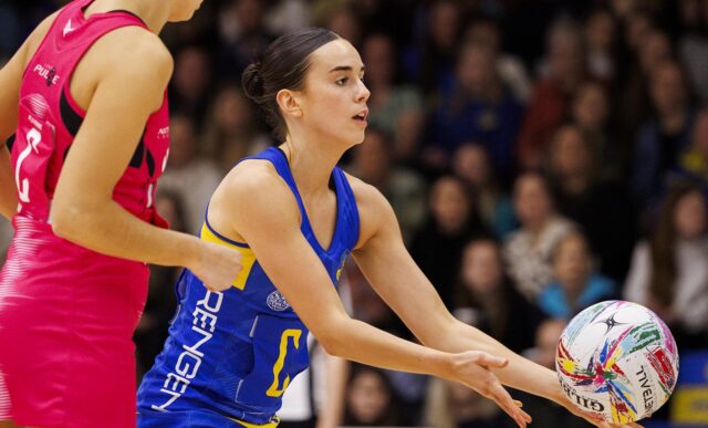 A photo of Team Bath Netball player Phoebe Maslen offloading the ball from her hands while under pressure from a London Pulse opponent, in front of a stand of supporters at the Team Bath Arena.