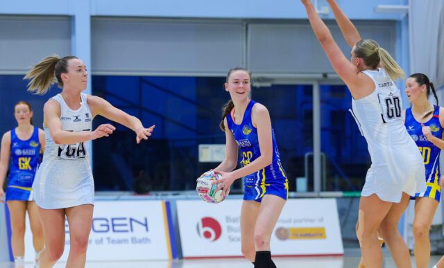 A photo of Team Bath Netball goal-attack holding the ball and smiling as she assesses her passing options while being closed down by two Birmingham Panthers defenders during a 2025 NXT Gen League fixture at the Team Bath Arena. The photo was taken by Josh Le Feuvre.