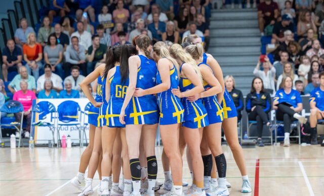 A photo of Team Bath Netball players doing a pre-match huddle in front of a stand of fans ahead of the NXT Gen League match against Leeds Rhinos on Sunday 15th June 2025.