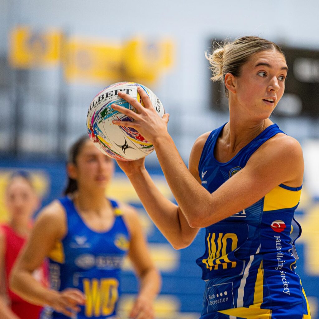 Team Bath Netball wing-attack Saskia Lea with the ball in both hands, turning to look down the court. Behind her in the background is team-mate Ellie Ervine. 
