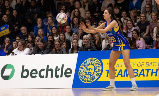 Team Bath Netball defender Orla Farrelly passes the ball in front of advertising boards featuring Bechtle and Team Bath branding.