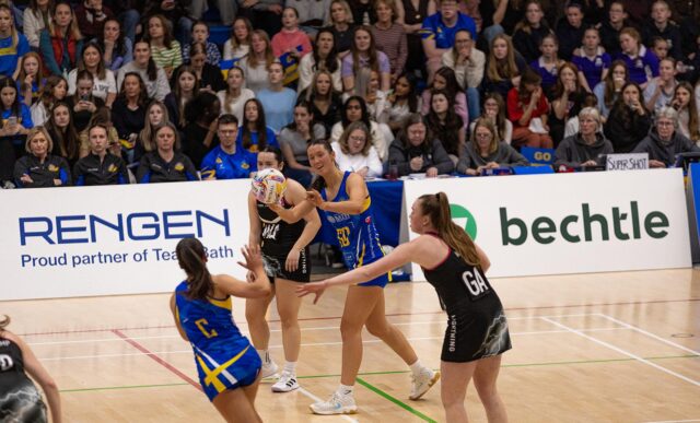 Team Bath Netball's Lottie Robinson shapes to pass the ball to team-mate Ellie Ervine as she is closed down by Loughborough Lightning defenders. Behind them is a stand full of spectators.