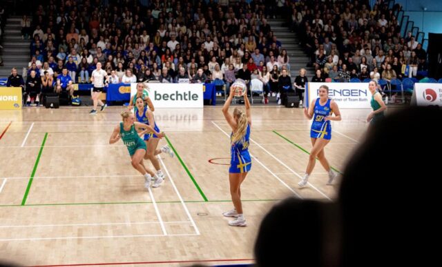 Team Bath Netball and LexisNexis Dragons players on court at the Team Bath Arena, as seen from the stands. Behind the players is another full stand of spectators.
