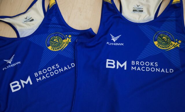 Two blue Team Bath Netball training tops on a table. Both have a Brooks Macdonald and Flyhawk logos on the front, plus the Team Bath Netball badge.